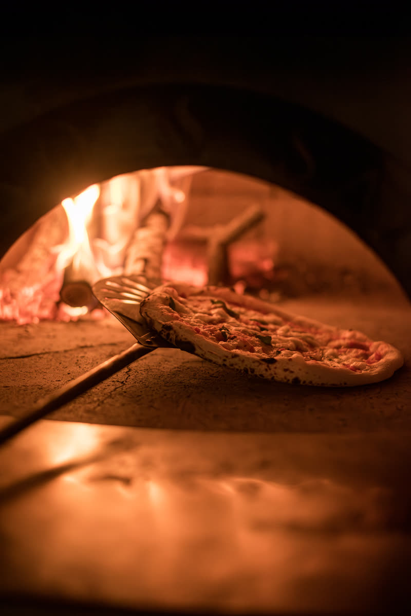 Uma pizza sendo retirada do forno na pá, com a brasa ao fundo.